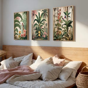 Three framed botanical prints depicting tropical scenes with green foliage, pink flowers, and birds. The artwork is set against a light background, creating a serene aesthetic. The frames are a light wood colour. The prints are in a bedroom setting.