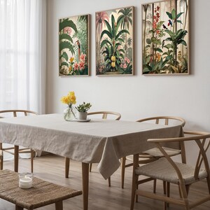 Three framed botanical prints with tropical plants and birds adorn a dining room wall. A beige linen tablecloth covers the table, surrounded by wooden chairs. A vase of yellow flowers and a candle add to the room's decor. The room has a natural feel.