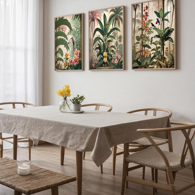 Three framed botanical prints with tropical plants and birds adorn a dining room wall. A beige linen tablecloth covers the table, surrounded by wooden chairs. A vase of yellow flowers and a candle add to the room's decor. The room has a natural feel.