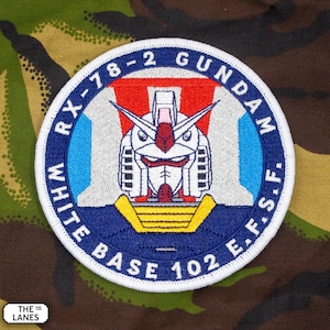 Może przedstawiać: Haftowana naszywka z motywem twarzy robota i napisem "RX-78-2 GUNDAM WHITE BASE 102 E.F.S.F." w okrągłym układzie. Naszywka ma niebieską obwódkę z białymi literami oraz czerwono-biało-niebieski wzór robota.