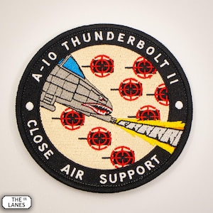 Op de afbeelding: Een geborduurde patch met een A-10 Thunderbolt II vliegtuig dat een kanon afvuurt op een doel met rode en zwarte bullseyes. De patch heeft een zwarte achtergrond met witte tekst die "A-10 THUNDERBOLT II CLOSE AIR SUPPORT" luidt.