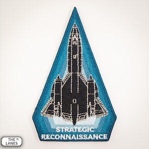 Könnte beinhalten: Ein blauer und weißer gestickter Aufnäher mit einer schwarzen Silhouette eines SR-71 Blackbird-Flugzeugs. Der Aufnäher hat die Form eines Dreiecks und trägt den Text "Strategic Reconnaissance" am unteren Rand.