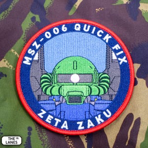 May include: A circular embroidered patch featuring a green robotic head with a single glowing white eye, set against a light blue background. The dark blue border contains the white text MSZ-006 QUICK FIX and ZETA ZAKU, finished with a red edge.