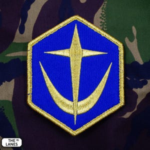 May include: A blue and gold embroidered patch featuring a stylised cross and crescent moon design. The patch is hexagonal in shape and has a gold border.