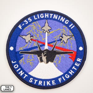 May include: A blue and white embroidered patch with the text "F-35 Lightning II Joint Strike Fighter". The patch features a black F-35 fighter jet with three smaller jets flying in formation.