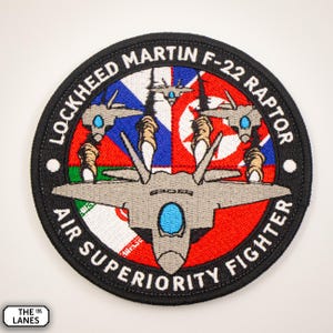 May include: A circular embroidered patch featuring a grey F-22 Raptor fighter jet in the centre. The jet is surrounded by the flags of Russia, North Korea, Iran, and the United Kingdom. The text "Lockheed Martin F-22 Raptor Air Superiority Fighter" is around the edge of the patch.