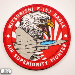 Könnte beinhalten: Ein gestickter Aufnäher in Rot, Weiß und Schwarz mit einem weißen Adlerkopf und einem grauen F-15J-Kampfjet. Der Text "MITSUBISHI F-15J EAGLE AIR SUPERIORITY FIGHTER" ist um den Rand des Aufnähers herum geschrieben.