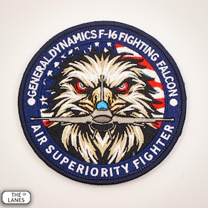 May include: A blue and white embroidered patch featuring an eagle's head and the text "General Dynamics F-16 Fighting Falcon Air Superiority Fighter".