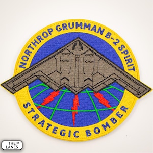 May include: A yellow, blue, and green embroidered patch with the text "Northrop Grumman B-2 Spirit Strategic Bomber". The patch features a gray silhouette of a B-2 bomber flying over a stylized globe.