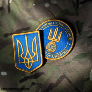 Könnte beinhalten: Zwei gestickte Patches, einer mit dem ukrainischen Dreizack in Gold und Blau, und der andere mit dem Text "Internationale Legion der Ukraine" in Gold auf blauem Hintergrund.