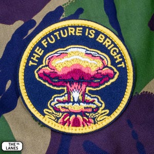May include: An embroidered patch in navy blue with a yellow border. The patch depicts a stylised mushroom cloud with the text "THE FUTURE IS BRIGHT" around the edge.