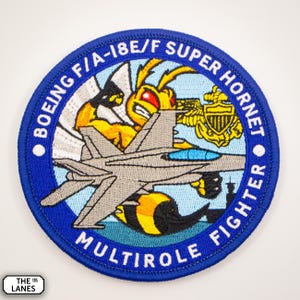 May include: A blue and white embroidered patch with the text "BOEING F/A-18E/F SUPER HORNET MULTIROLE FIGHTER" around the edge. A cartoon hornet with a jet fighter is in the centre of the patch.