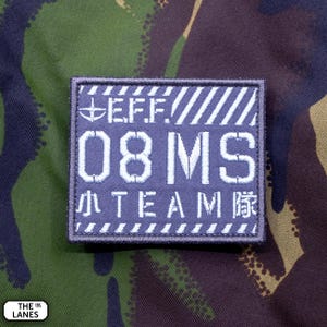 Pode incluir: Um emblema retangular bordado em azul escuro com texto e gráficos brancos. Apresenta as letras E.F.F., o número 08MS e a palavra TEAM ladeada por caracteres japoneses. O design inclui listras brancas diagonais na parte superior e inferior.