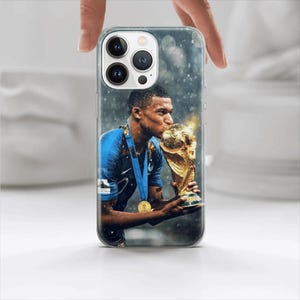 Puede incluir: Una funda transparente para teléfono con una foto de un jugador de fútbol besando un trofeo dorado. El jugador lleva una camiseta azul con una medalla de oro alrededor del cuello. El fondo es una imagen borrosa de un campo nevado.