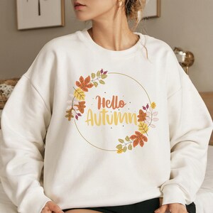 Könnte beinhalten: Cremefarbenes Sweatshirt mit einem kreisförmigen Blumendesign mit Herbstblättern in Orange, Gelb und Braun. Die Worte "Hello Autumn" sind in einer Schriftart innerhalb des Kreises geschrieben. Ein gemütliches Kleidungsstück zum Thema Herbst.