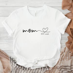 Könnte beinhalten: Weißes T-Shirt mit dem Wort "Mom" in Schreibschrift, gefolgt von einem Herz und dem Text "EST. 2026" in Schwarz. Das Shirt besteht aus einem weichen Material und hat kurze Ärmel.