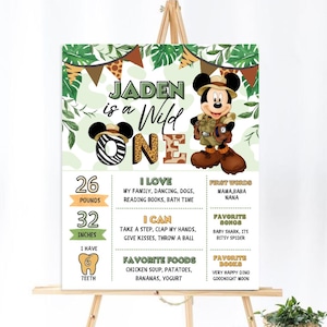 May include: A green and brown jungle-themed first birthday milestone poster featuring a cartoon Mickey Mouse dressed as an explorer. The poster includes the name "Jaden" and the text "is a Wild One". It also lists the child's age, weight, height, favorite foods, books, and first words.