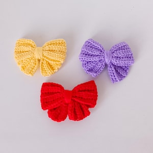 May include: Three handmade crochet bows in yellow, purple, and red. The bows are arranged on a white surface. Each bow has a textured, ribbed appearance, with a classic bow shape. These bows are suitable for hair accessories or craft projects.
