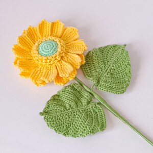 May include: A crocheted sunflower with yellow petals and a green centre, with two green crocheted leaves attached to a brown stem.
