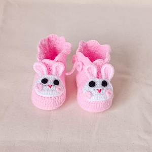 Könnte beinhalten: Ein Paar rosa gehäkelte Babyschuhe. Jeder Schuh hat ein Hasengesicht mit schwarzen Augen, einer weißen Schnauze und einem roten Mund. Die Schuhe haben einen gerüschten oberen Rand und sind so konzipiert, dass sie die Füße des Babys warm halten.