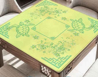 May include: A square light yellow game mat featuring green sea turtle illustrations, floral patterns, and geometric corner borders. The mat is displayed on a wooden table with four chairs, accompanied by insets showing playing cards and tiles.