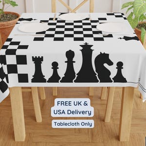 Puede incluir: Mantel blanco con un diseño de tablero de ajedrez en blanco y negro y siluetas de piezas de ajedrez negras. El mantel está sobre una mesa de madera, con platos blancos. El texto "Tablecloth Only" es visible.