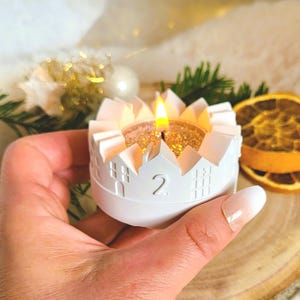 Könnte beinhalten: Ein weißer Kerzenhalter mit hausförmigem Design und der Zahl 2. Der Kerzenhalter hat eine brennende Kerze mit goldglitzerndem Innenraum. Der Kerzenhalter wird in einer Hand gehalten, mit getrockneten Orangenscheiben und Grünzeug im Hintergrund.