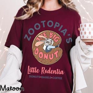 Puede incluir: Camiseta color granate con una imagen de dibujos animados de Judy Hopps de Zootopia dentro de un donut. El texto dice "Zootopia's The Big Donut" y "Little Rodentia". Debajo del texto est&aacute;n las palabras "DONUTS &bull; BAGELS &bull; COFFEE".