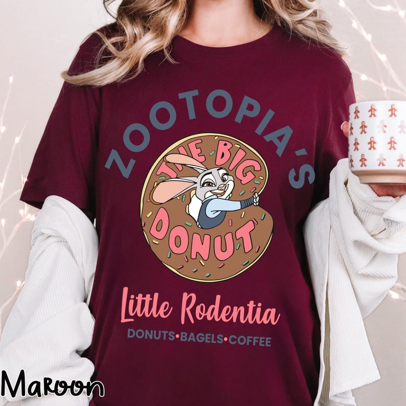 Puede incluir: Camiseta color granate con una imagen de dibujos animados de Judy Hopps de Zootopia dentro de un donut. El texto dice "Zootopia's The Big Donut" y "Little Rodentia". Debajo del texto est&aacute;n las palabras "DONUTS &bull; BAGELS &bull; COFFEE".