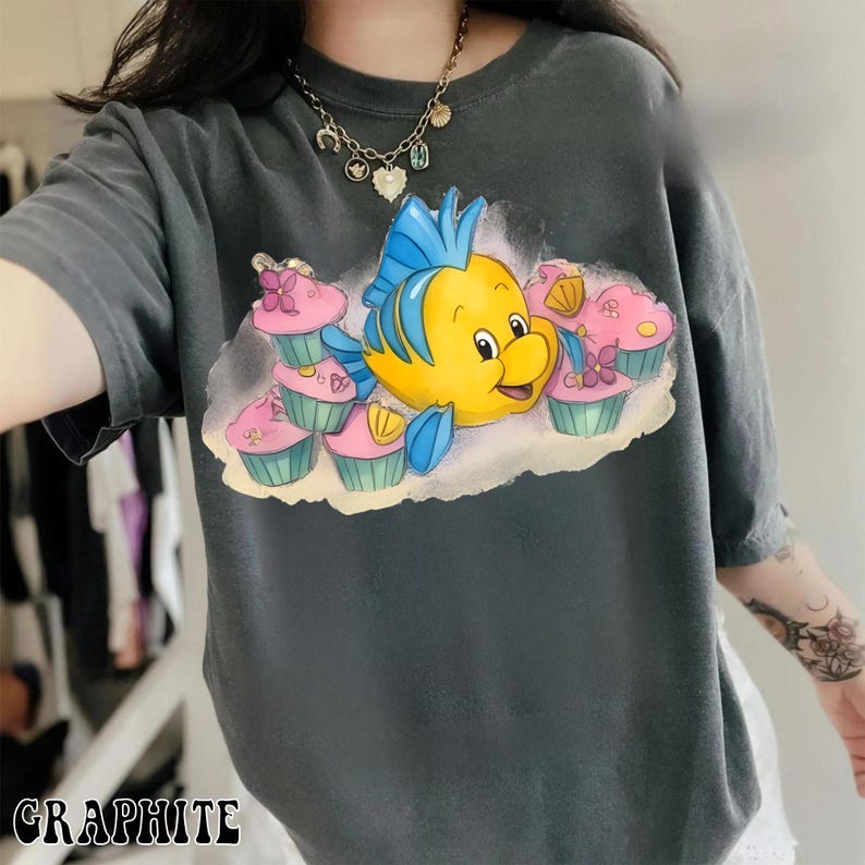 May include: Graphite gray t-shirt featuring a cartoon illustration of a yellow fish with blue fins and a smiling face, surrounded by pink cupcakes. The word "GRAPHITE" is printed at the bottom of the shirt.