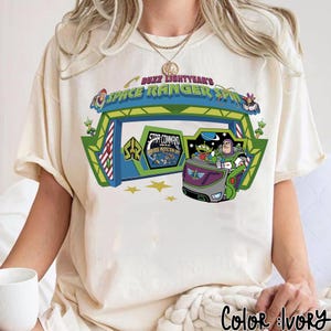 K&ouml;nnte beinhalten: Elfenbeinfarbenes T-Shirt mit einer farbenfrohen Grafik von Buzz Lightyear's Space Ranger Spin. Das Design beinhaltet das Star Command-Logo, Aliens und Buzz Lightyear in einem Fahrzeug. Der Text "Buzz Lightyear's Space Ranger Spin" ist oben gew&ouml;lbt.
