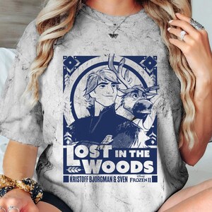 Puede incluir: Una camiseta gris te&ntilde;ida con un dise&ntilde;o azul de Frozen II de Disney. El dise&ntilde;o incluye el texto "LOST IN THE WOODS" con im&aacute;genes de Kristoff, Sven y elementos decorativos. La camiseta tiene cuello redondo.