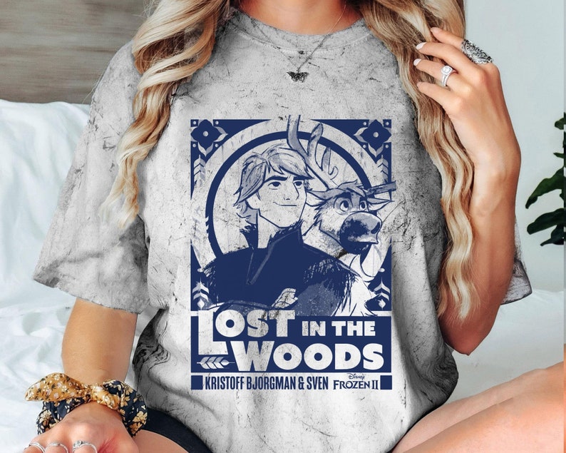 Puede incluir: Una camiseta gris te&ntilde;ida con un dise&ntilde;o azul de Frozen II de Disney. El dise&ntilde;o incluye el texto "LOST IN THE WOODS" con im&aacute;genes de Kristoff, Sven y elementos decorativos. La camiseta tiene cuello redondo.