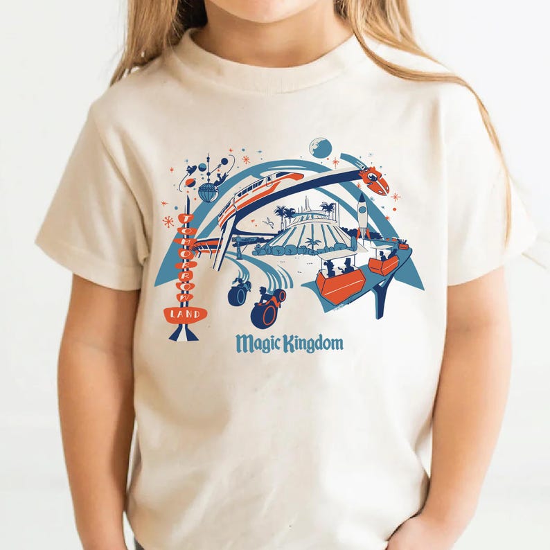 Op de afbeelding: Een cr&egrave;mekleurig T-shirt met een kleurrijke afbeelding van een themaparksc&egrave;ne. Het ontwerp bevat een achtbaan, een raket en andere attracties, met de woorden "LAND" en "Magic Kingdom" in blauw en oranje.
