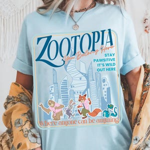 Puede incluir: Camiseta azul claro con un dise&ntilde;o de Zootopia. El dise&ntilde;o incluye la palabra "Zootopia" en azul, personajes de dibujos animados y la frase "Where anyone can be anything".
