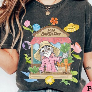 Op de afbeelding: Donkergrijze t-shirt met een cartoonkonijn met een strohoed en een roze geruit overhemd, die een wortel in een tuin vasthoudt. Het ontwerp bevat de tekst "2026 Earth Day" en bloemmotieven. Er is ook een gele hoed met "2026".
