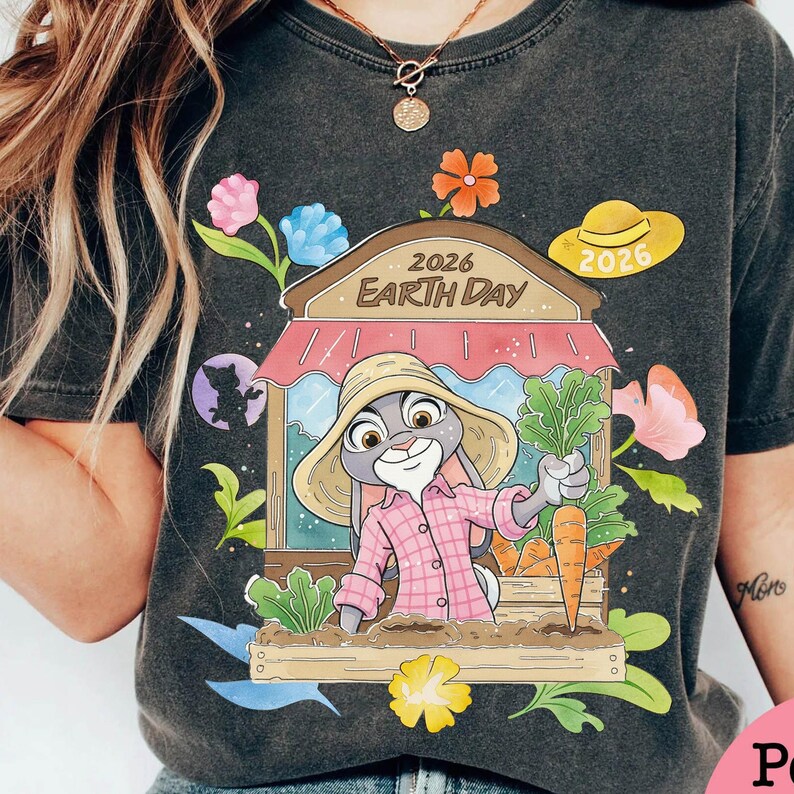 Op de afbeelding: Donkergrijze t-shirt met een cartoonkonijn met een strohoed en een roze geruit overhemd, die een wortel in een tuin vasthoudt. Het ontwerp bevat de tekst "2026 Earth Day" en bloemmotieven. Er is ook een gele hoed met "2026".