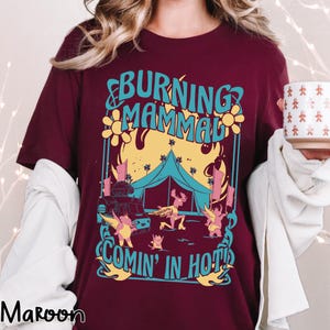 Puede incluir: Camiseta granate con un dise&ntilde;o gr&aacute;fico colorido. El dise&ntilde;o presenta el texto "BURNING MAMMAL COMIN' IN HOT!" con ilustraciones de una tienda de campa&ntilde;a, un cami&oacute;n de comida y criaturas aladas. La camiseta es de manga corta.
