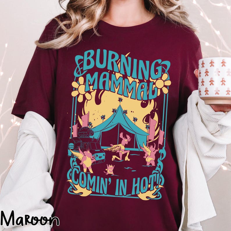 Puede incluir: Camiseta granate con un dise&ntilde;o gr&aacute;fico colorido. El dise&ntilde;o presenta el texto "BURNING MAMMAL COMIN' IN HOT!" con ilustraciones de una tienda de campa&ntilde;a, un cami&oacute;n de comida y criaturas aladas. La camiseta es de manga corta.