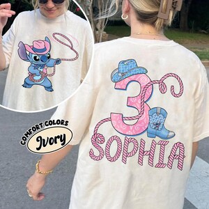 May include: Ivory-colored t-shirt featuring a cartoon Stitch character in a cowboy hat, roping. The back of the shirt displays a pink number 3, a cowboy hat, a blue boot, and the name "Sophia" in pink rope lettering. The shirt is from Comfort Colors.
