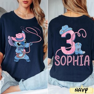 May include: Navy blue t-shirt featuring a cartoon character wearing a pink cowboy hat and roping. The back of the shirt displays the number "3", a cowboy boot, a cowboy hat, and the name "SOPHIA" in pink.