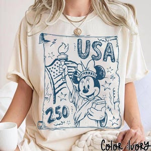 May include: Ivory-colored t-shirt featuring a blue graphic of Minnie Mouse as the Statue of Liberty, holding a torch and a book, with the word "USA" above. The design includes the American flag and stars.