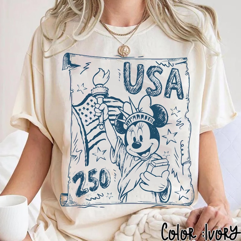 May include: Ivory-colored t-shirt featuring a blue graphic of Minnie Mouse as the Statue of Liberty, holding a torch and a book, with the word "USA" above. The design includes the American flag and stars.
