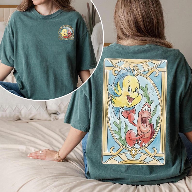 May include: Teal-colored t-shirt featuring a colorful illustration of Flounder and Sebastian from The Little Mermaid on the back. The front of the shirt has a smaller graphic of Ariel and Flounder. The shirt has a crew neck and short sleeves.