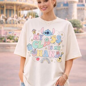 Puede incluir: Camiseta color crema con el texto "My Class is My Ohana" en letras coloridas de estilo caricatura. El dise&ntilde;o incluye un extraterrestre azul, un libro, un arco&iacute;ris y otros elementos l&uacute;dicos. La camiseta la lleva una persona.