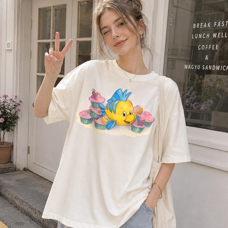 May include: Cream-colored t-shirt featuring a cartoon illustration of a yellow fish with blue fins and pink cupcakes. The short-sleeved shirt is made of a soft material and is suitable for casual wear.
