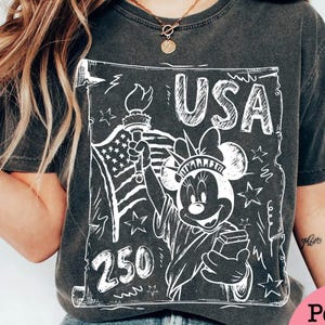 May include: Dark gray t-shirt featuring a white graphic of Minnie Mouse as the Statue of Liberty, holding a torch and a book. The graphic includes the text "USA" and the number "250", with the American flag in the background.