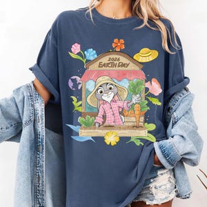 Op de afbeelding: Marineblauw T-shirt met een cartoonkonijn in een roze geruit overhemd en strohoed, met wortels. Het ontwerp bevat een winkelgevel met de tekst "2026 Earth Day", omringd door bloemen en bladeren. Een gele zonnehoed is ook inbegrepen.
