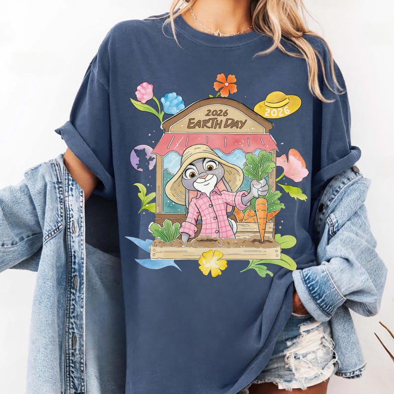 Op de afbeelding: Marineblauw T-shirt met een cartoonkonijn in een roze geruit overhemd en strohoed, met wortels. Het ontwerp bevat een winkelgevel met de tekst "2026 Earth Day", omringd door bloemen en bladeren. Een gele zonnehoed is ook inbegrepen.