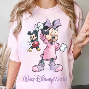 May include: A light pink t-shirt featuring a cartoon Minnie Mouse character holding a Mickey Mouse character. Minnie is wearing a pink dress and bow. The text "Walt Disney World" is printed in a cursive font at the bottom of the shirt.